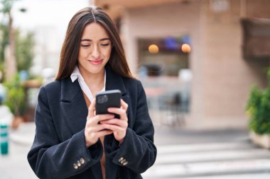 Genç, güzel, İspanyol bir kadın sokakta akıllı telefon kullanarak gülümsüyor.