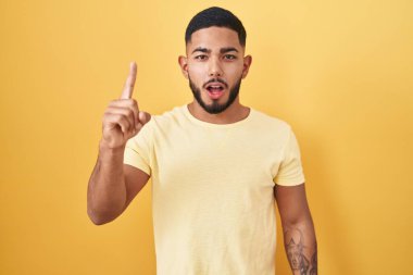 Young hispanic man standing over yellow background pointing finger up with successful idea. exited and happy. number one. 