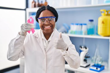 African american woman working at scientist laboratory holding pink ribbon smiling happy and positive, thumb up doing excellent and approval sign 