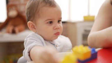 Adorable toddler holding plastic construction blocks sitting on table at kindergarten