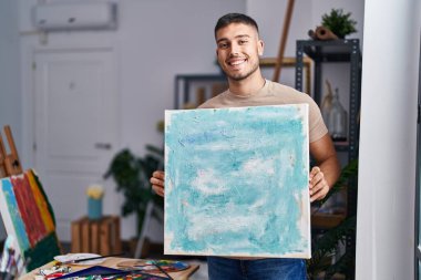 Young hispanic painter man holding canvas smiling with a happy and cool smile on face. showing teeth. 