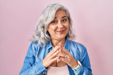 Middle age woman with grey hair standing over pink background hands together and fingers crossed smiling relaxed and cheerful. success and optimistic 