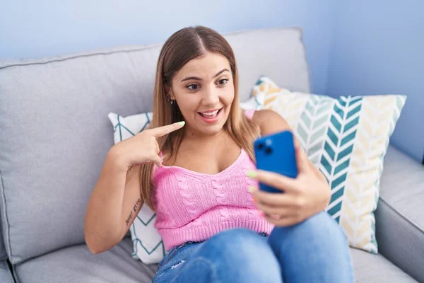 年轻的金发女人拿着智能手机在沙发上做视频通话 开心地微笑着 用手和手指指指点点 — 图库照片