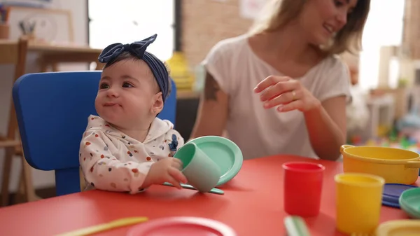 Woman and toddler learning to eat with plastic dish toy sitting on table at kindergarten