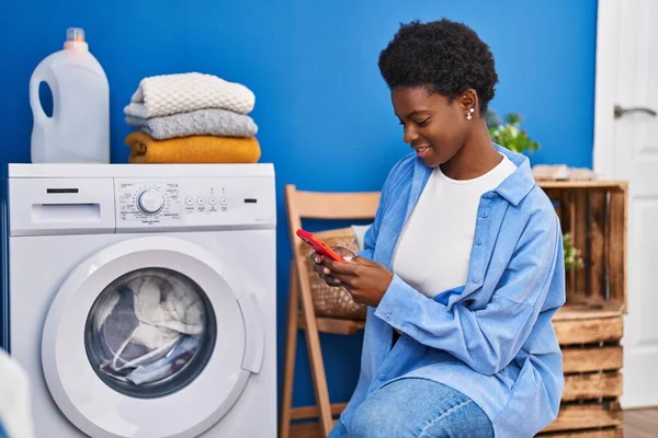 在洗衣房用智能手机等洗衣机的非洲裔美国女人 — 图库照片