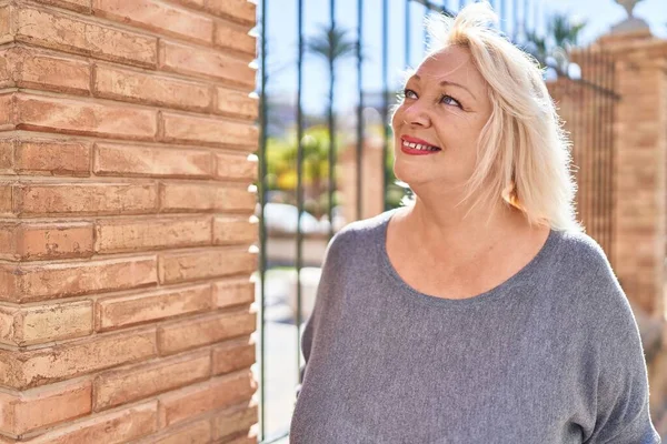 Orta yaşlı sarışın kadın sokakta kendinden emin bir şekilde gülümsüyor.