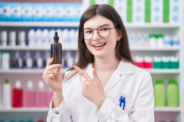 Young caucasian woman working at pharmacy drugstore holding serum smiling happy pointing with hand and finger