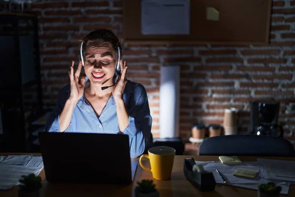 Beautiful brunette woman working at the office at night covering ears with fingers with annoyed expression for the noise of loud music. deaf concept.