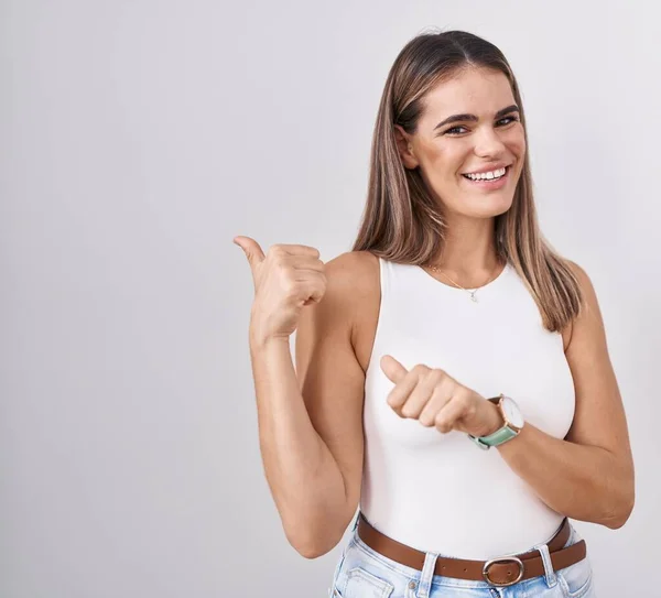 Hispanic young woman standing over white background pointing to the back behind with hand and thumbs up, smiling confident