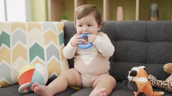 Adorable toddler bitting plastic hoop sitting on sofa at home