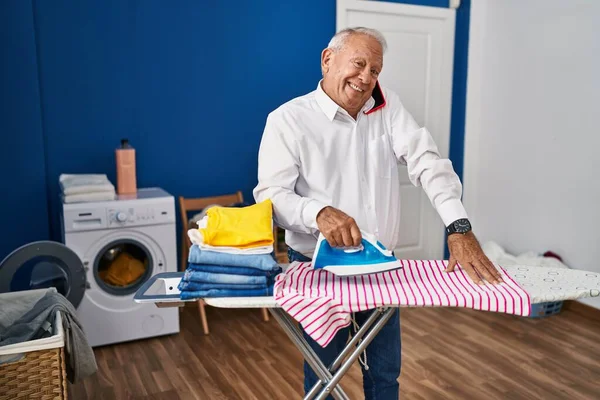 Senior man talking on the smartphone ironing clothes at laundry room