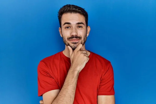 Young hispanic man with beard wearing red t shirt over blue background looking confident at the camera with smile with crossed arms and hand raised on chin. thinking positive.