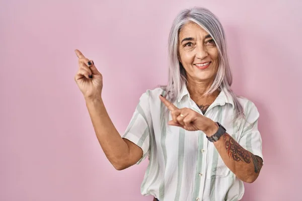 Middle age woman with grey hair standing over pink background smiling and looking at the camera pointing with two hands and fingers to the side.
