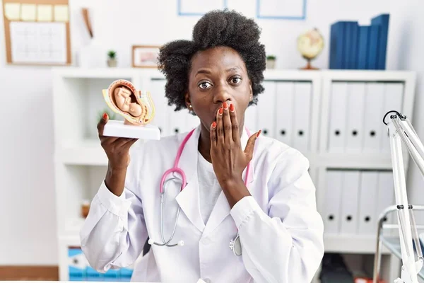 African doctor woman holding anatomical model of female uterus with fetus covering mouth with hand, shocked and afraid for mistake. surprised expression