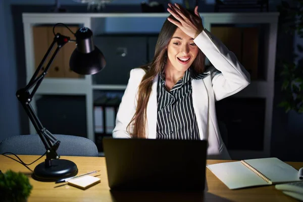 Young brunette woman working at the office at night with laptop surprised with hand on head for mistake, remember error. forgot, bad memory concept.