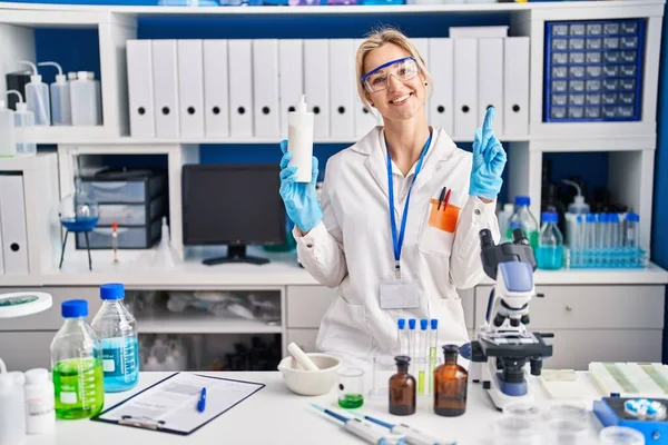 Young caucasian woman working at scientist laboratory holding body lotion smiling with an idea or question pointing finger with happy face, number one