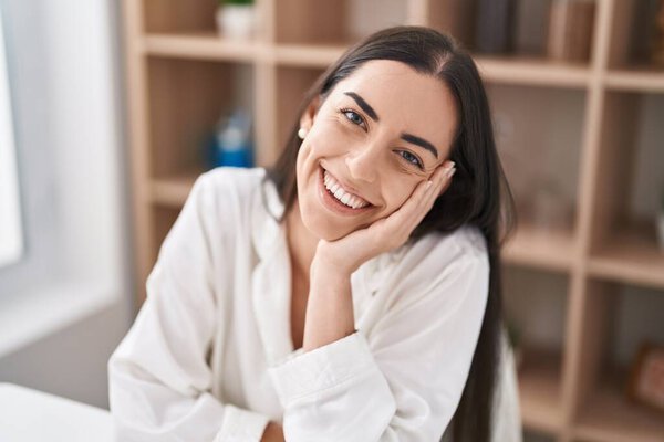 Молодая латиноамериканка, уверенно улыбающаяся, сидя дома на столе