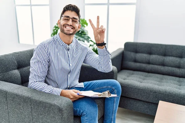Young psychologist man at consultation office smiling with happy face winking at the camera doing victory sign with fingers. number two.