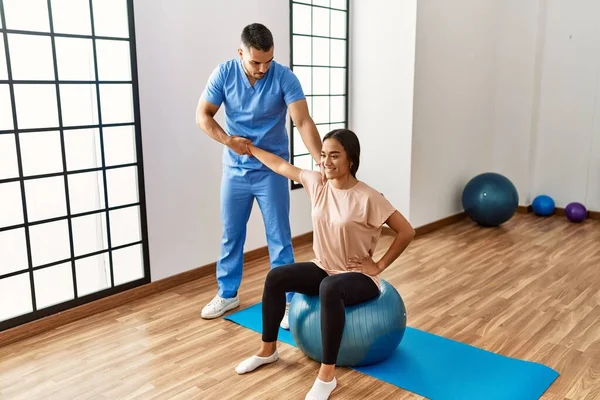 Latin man and woman wearing physiotherapist uniform having rehab session using fit ball at rehab center