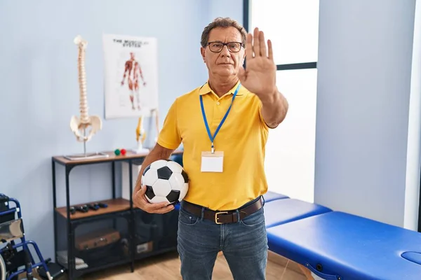 Senior man working at sport physiotherapy clinic with open hand doing stop sign with serious and confident expression, defense gesture