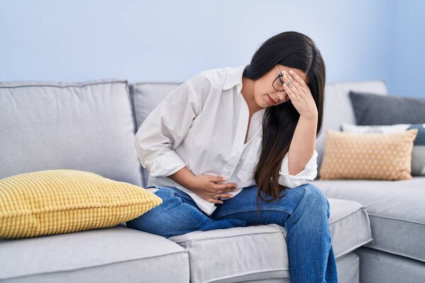 Young hispanic woman sitting on sofa suffering for stomach pain at home
