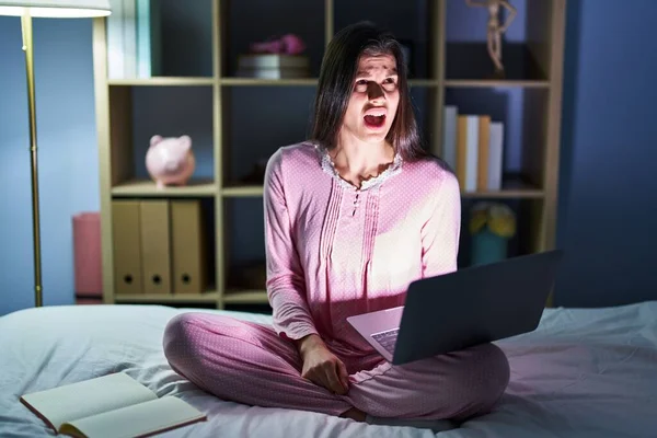 Young hispanic woman using computer laptop on the bed angry and mad screaming frustrated and furious, shouting with anger. rage and aggressive concept.