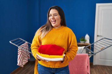 Young hispanic woman holding clean and folded laundry winking looking at the camera with sexy expression, cheerful and happy face. 