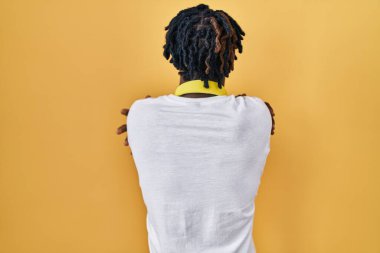 Young african man with dreadlocks standing over yellow background hugging oneself happy and positive from backwards. self love and self care 
