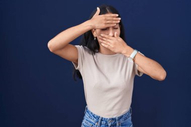 Mavi arka planda duran genç İspanyol kadın gözlerini ve ağzını elleriyle kapatıyor, şaşırmış ve şok olmuş. duygu gizleme 