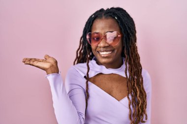 African woman with braided hair standing over pink background smiling cheerful presenting and pointing with palm of hand looking at the camera. 