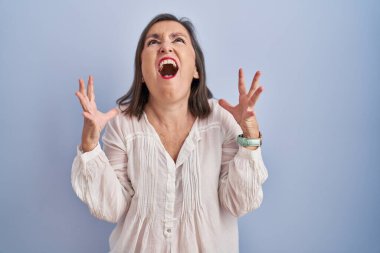 Middle age hispanic woman standing over blue background crazy and mad shouting and yelling with aggressive expression and arms raised. frustration concept. 