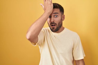 Handsome hispanic man standing over yellow background surprised with hand on head for mistake, remember error. forgot, bad memory concept. 