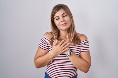 Soyutlanmış arka planda duran genç İspanyol kadın elleri göğsünde, gözleri kapalı ve yüzünde minnet dolu bir jest ile gülümsüyor. sağlık kavramı. 