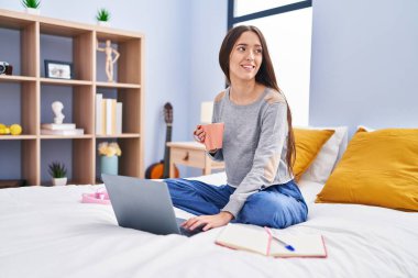Young beautiful hispanic woman student drinking coffee studying on bed at bedroom