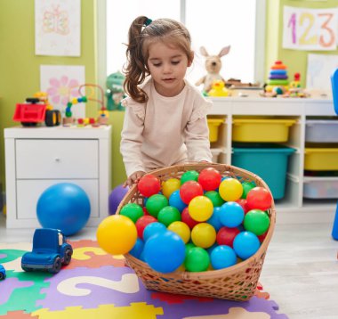 Adorable hispanic girl playing with balls standing at kindergarten