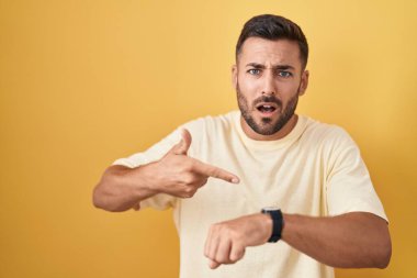 Handsome hispanic man standing over yellow background in hurry pointing to watch time, impatience, upset and angry for deadline delay 
