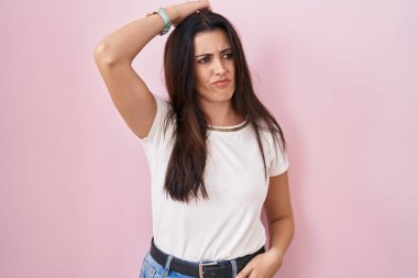 Young brunette woman standing over pink background confuse and wondering about question. uncertain with doubt, thinking with hand on head. pensive concept. 
