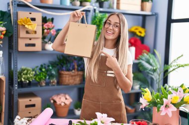 Young blonde woman working at florist shop holding bag smiling happy pointing with hand and finger 