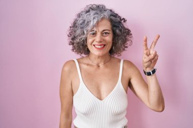 Pembe arka planda duran gri saçlı orta yaşlı bir kadın iki numaralı parmaklarıyla gülümseyerek kendinden emin ve mutlu bir şekilde onu işaret ediyor.. 