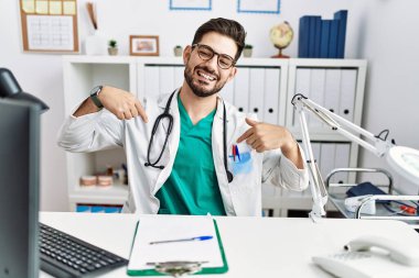 Young man with beard wearing doctor uniform and stethoscope at the clinic looking confident with smile on face, pointing oneself with fingers proud and happy. 