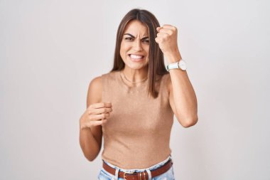 Young hispanic woman standing over white background angry and mad raising fist frustrated and furious while shouting with anger. rage and aggressive concept. 