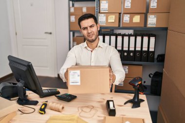 Young hispanic man with beard working at small business ecommerce holding box clueless and confused expression. doubt concept. 