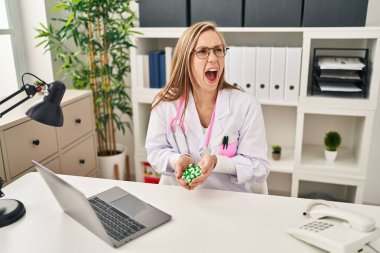 Young blonde doctor woman holding pills angry and mad screaming frustrated and furious, shouting with anger. rage and aggressive concept. 
