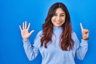 Hispanic young woman standing over blue background showing and pointing up with fingers number six while smiling confident and happy. 