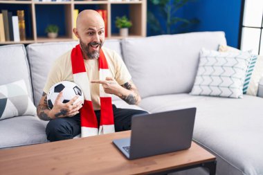 Hispanic man with tattoos watching football match hooligan holding ball on the laptop smiling happy pointing with hand and finger 
