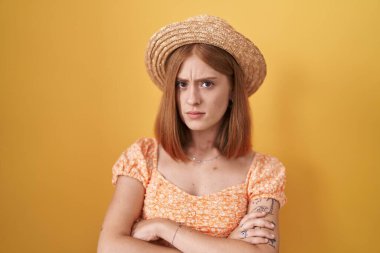 Young redhead woman standing over yellow background wearing summer hat skeptic and nervous, disapproving expression on face with crossed arms. negative person. 