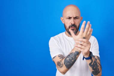 Hispanic man with tattoos standing over blue background suffering pain on hands and fingers, arthritis inflammation 