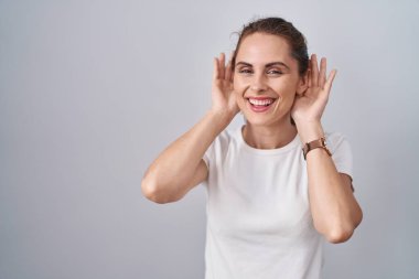 Beautiful brunette woman standing over isolated background trying to hear both hands on ear gesture, curious for gossip. hearing problem, deaf 