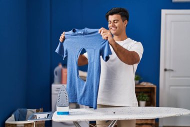 Hispanic man ironing holding burned iron shirt at laundry room smiling with a happy and cool smile on face. showing teeth. 