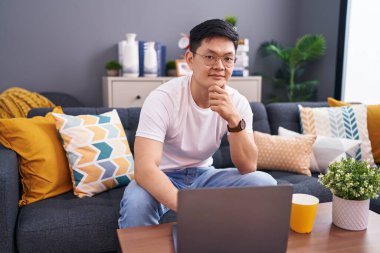 Young asian man using laptop at home sitting on the sofa looking confident at the camera smiling with crossed arms and hand raised on chin. thinking positive. 